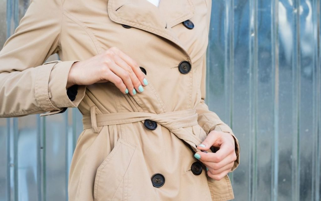 woman tying belt of classic brown trench coat blue manicure