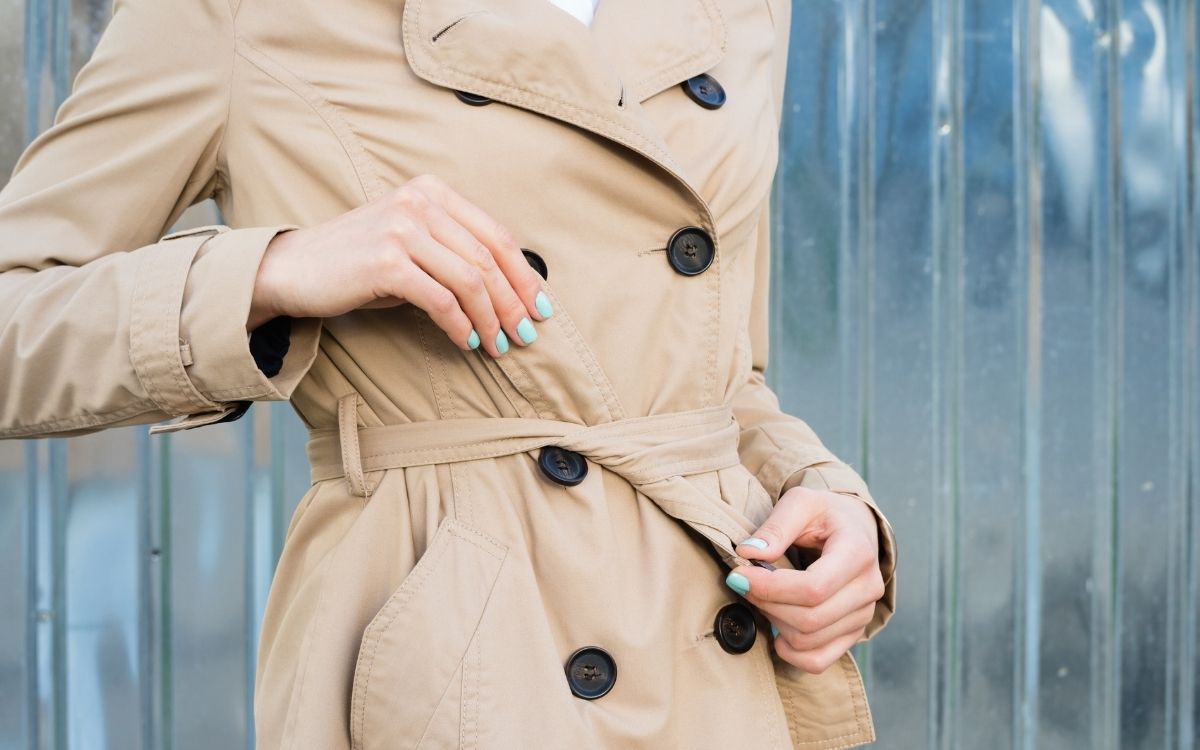 woman tying belt of classic brown trench coat blue manicure