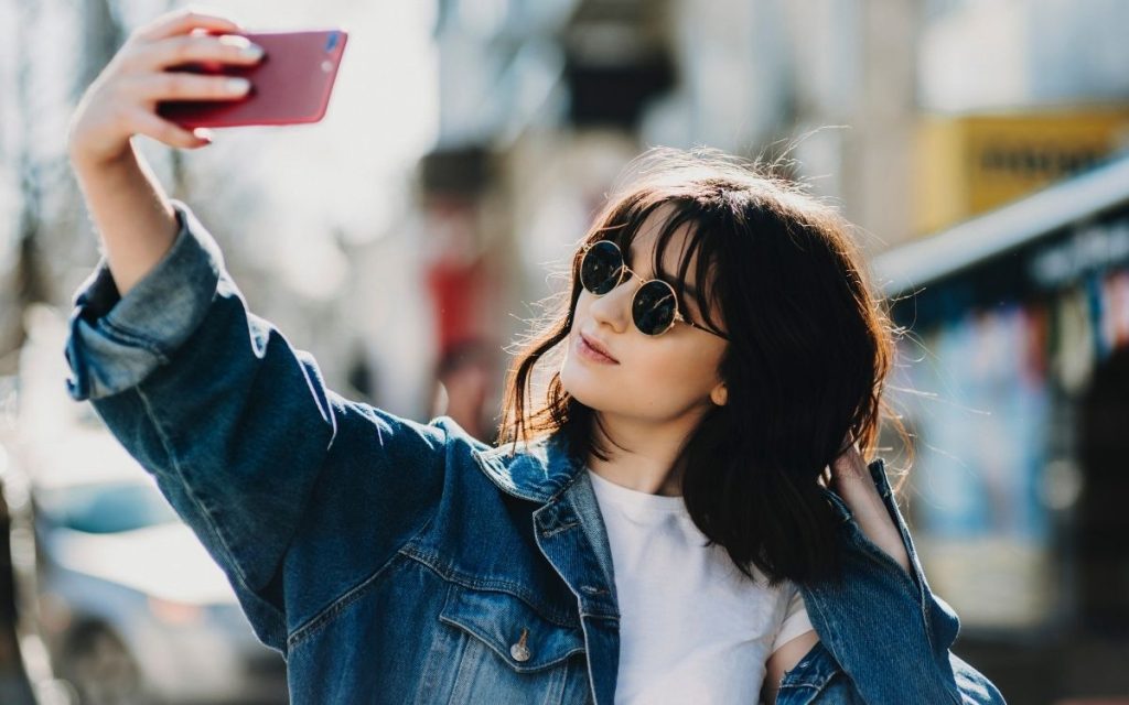 woman wearing sunglasses and oversized dark jean jacket denim jacket taking a selfie
